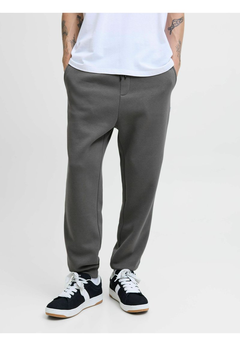 Grå sweatpants med en afslappet pasform, der har sidelommer og snoretræk i taljen, parret med sorte og hvide sports sneakers.