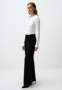 Haut ribbed blanc à manches longues associé à un pantalon noir taille haute et des escarpins noirs à bouts pointus, sur un fond blanc uni.