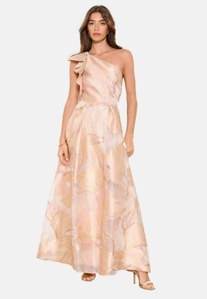 Frau, die ein bodenlanges, einseitig schulterfreies pfirsichfarbenes Kleid mit Rüschendetail und subtilem Blumenmuster trägt, kombiniert mit nudefarbenen Sandalen mit hohen Absätzen.