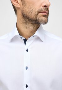 Camicia bianca con colletto classico, dettagli a pois blu navy all'interno del colletto e bottoni blu, realizzata in un tessuto liscio e strutturato.