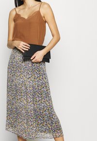 Top in raso marrone con pizzo, abbinato a una gonna midi fluida a fantasia floreale in viola, giallo e nero. Tiene una pochette nera.