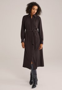 Robe marron à manches longues avec col, taille ceinturée et fente sur le côté. Tissu lisse avec un design structuré, associée à des bottes noires hautes jusqu'aux genoux.