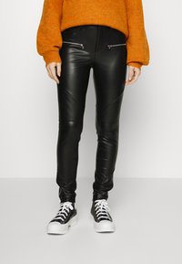 Calças de couro sintético pretas com detalhes diagonais e bolsos com fecho, combinadas com ténis em preto e branco e um suéter laranja.