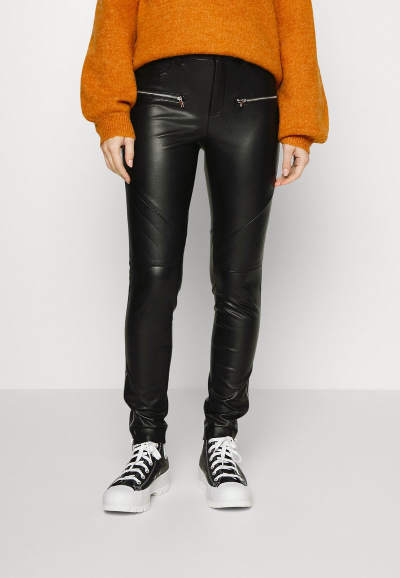 Calças de couro sintético pretas com detalhes diagonais e bolsos com fecho, combinadas com ténis em preto e branco e um suéter laranja.