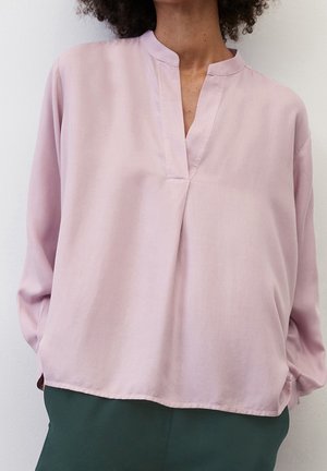 Kvinde iført en løs, lyserød bluse med lange ærmer og V-formet udskæring samt mørkegrønne bukser mod en ensartet hvid baggrund.