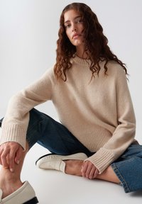 Giovane donna con capelli ricci seduta con le gambe incrociate, indossa un maglione beige, jeans blu e mocassini bianchi su uno sfondo bianco semplice.