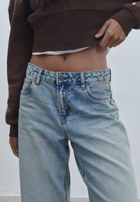 Svijetloplave traperice s visokim strukom, s istaknutim detaljima habanja i opuštenim krojem. Struk ima metalnu dugmad i petlje za remen.