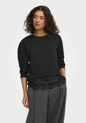Femme aux cheveux bouclés portant un pull noir et un pantalon gris à fines rayures avec un détail en dentelle, debout avec une main dans la poche sur fond blanc.