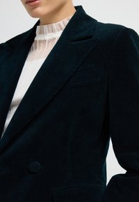 Detailní záběr na osobu v tmavě zeleném kordovém blejzru přes bílou průhlednou blůzu s vysokým límcem.