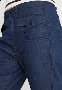 Detailní záběr na modré džíny z denimu s knoflíkovými klopovými kapsami a viditelným švem, nošené s pruhovaným topem.