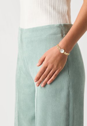 Light green corduroy pants with vertical ribbing, paired with a white ribbed top. A gold watch with a round face and decorative links on the wrist.