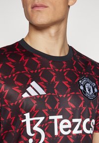Musta ja punainen Manchester Unitedin pelipaita kulmikkailla punaisilla kuvioilla, valkoinen Adidas-logo ja sponsorin teksti "tz Tezcs". Pyöreä kaula-aukko.