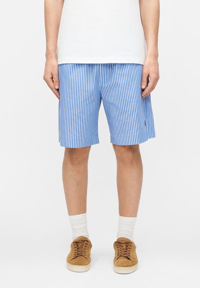 Homme portant un short à rayures verticales bleu et blanc, des chaussettes blanches, des baskets marron et une chemise blanche, debout devant un fond blanc.