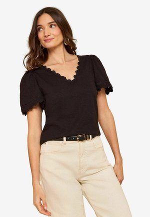 Hosszú barna hajú nő fekete, hullámos V-nyakú, rövid ujjú felsőben és világos bézs, magas derekú nadrágban, fekete övvel.