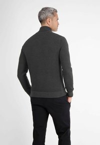 Mann mit grauem Haar, der einen dunkelgrauen strukturierten Pullover und schwarze Hosen trägt, mit Händen in den Taschen stehend, mit dem Rücken zu einer weißen Hintergrund.