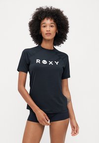 Zwarte rash guard met korte mouwen, gemaakt van een glad materiaal en met het woord "ROXY" in het wit op de voorkant gedrukt.