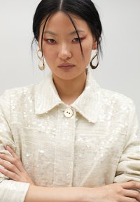 Vit paljettsjöja med krage och knappstängning, i texturerad tyg. Modellen har armarna i kors och bär guldörhängen.