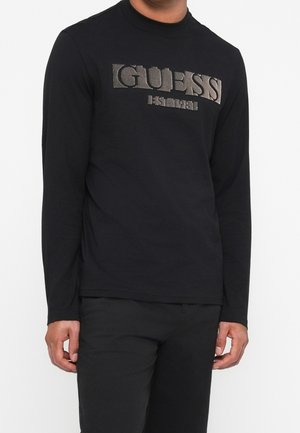 Mężczyzna w czarnej koszulce z długim rękawem z szarym logo "GUESS EST. 1981" na piersi i czarnych spodniach.