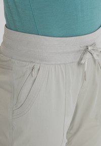 Jasnoszare spodnie z elastycznym pasem, sznurkiem do regulacji i zasuwaną boczną kieszenią, noszone z turkusową, prążkowaną bluzką widoczną w talii.