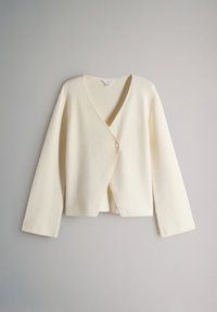 Cardigan en maille couleur crème à manches longues, col en V profond et fermeture par un bouton sur fond gris.