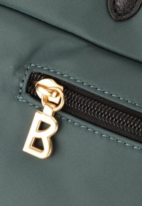 Detalle de una bolsa de tela verde con un zipper negro. Presenta un tirador del zipper dorado con forma de "B." Material texturizado con costuras visibles.