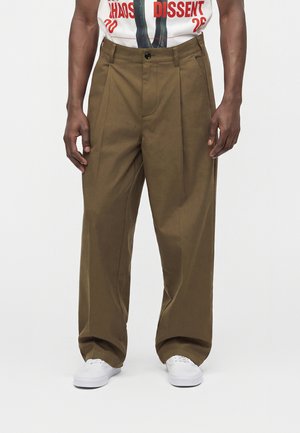 Mann trägt lockere braune Hose, weiße Sneakers und ein weißes T-Shirt mit rotem Text und steht vor einem schlichten Hintergrund.