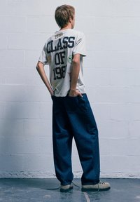 Młoda osoba w za dużych dżinsach i szarej koszulce z napisem "Klasa 1987" stoi przodem do białej ceglanej ściany z rękami w tylnej kieszeni.