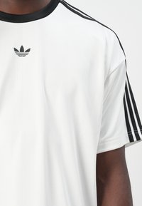 Witte sport t-shirt met zwarte accenten, een geribde kraag en drie zwarte strepen op de mouwen, met een klein zwart logo op de borst.