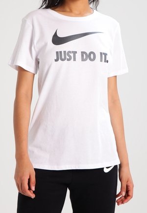 Persona vistiendo una camiseta blanca de Nike con el texto y el logo "JUST DO IT.", combinada con pantalones negros que tienen un pequeño logotipo de Nike.
