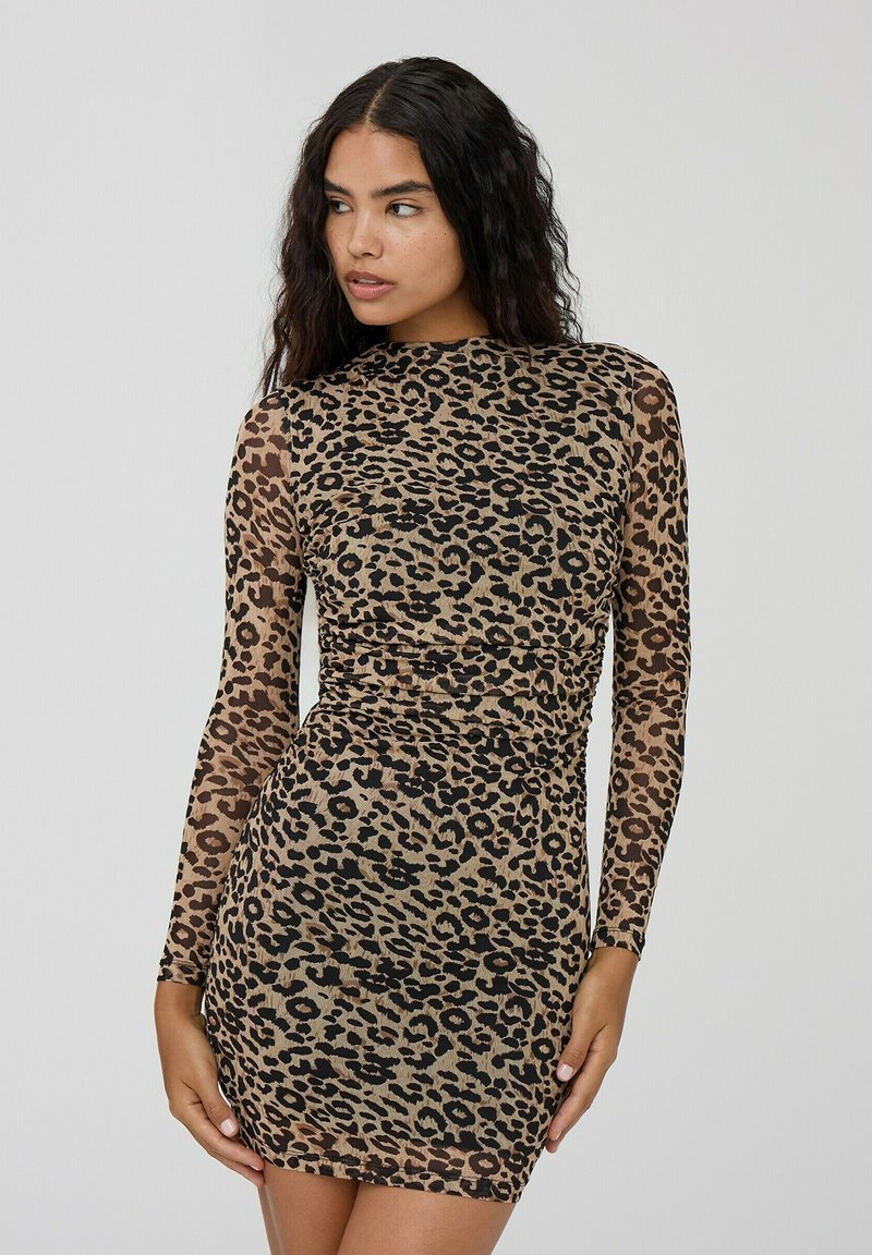 Donna con lunghi capelli ricci scuri che indossa un vestito aderente a maniche lunghe con stampa leopardata, in posa con le mani vicino alle cosce su uno sfondo semplice.
