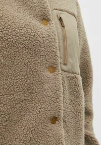 Giacca in sherpa fleece beige con bottoni a pressione color marrone e una tasca con zip sul petto con un tirazip dorato sul lato destro.