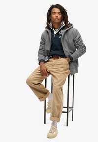 Grijze rits-hoodie over een marineblauwe polo, beige chino's en witte sneakers, zittend op een zwart metalen kruk met een neutrale achtergrond.