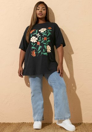 Kobieta w czarnej, dużej koszulce z dużym kwiatowym nadrukiem, jasno-niebieskich dżinsach i białych trampkach, stojąca na tle beżowej, teksturowanej ściany.