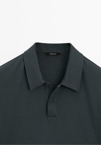 Polo vert foncé avec col et deux boutons-pression visibles, affichant une étiquette Massimo Dutti à l'intérieur de l'encolure.