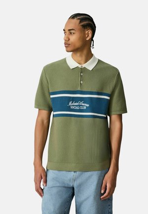 Junger Mann mit geflochtenem Haar, der ein olivgrünes gestricktes Polo mit blau-weißem Streifen und dem Schriftzug „Melrose Avenue Social Club“ trägt, kombiniert mit hellen Jeans.