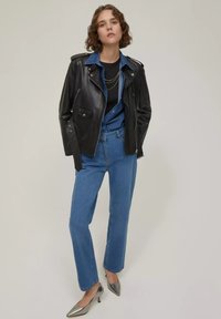 Donna con capelli corti e ricci che indossa una giacca di pelle nera sopra una camicia di denim blu, jeans blu e tacchi a punta metallici, in piedi su uno sfondo neutro.