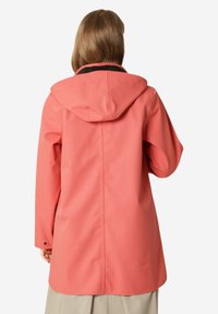Impermeable de color coral con capucha, fabricado con un material suave y resistente al agua. Presenta mangas largas y un dobladillo trasero ligeramente más largo. Detalles de ribete negro.