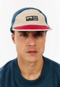 Young man wearing a blue and beige cap with red brim and “Pro Test” logo, and a dark blue crew neck sweater.