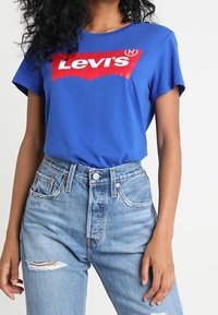 Blå bomullströja med en framträdande röd Levi's-logotyp, kombinerad med högmidjade ljusblå denimjeans med sliten detalj.