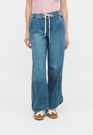 Personne portant un pantalon en denim bleu à jambes larges avec taille élastique et cordon blanc, assorti à des baskets marron et blanches.