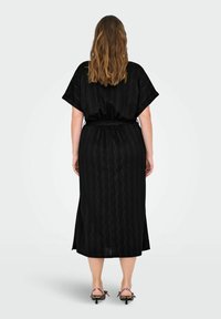 Črna, obleka srednje dolžine z valovito teksturo, ohlapnimi rokavi in pasom za zavezovanje v pasu. Manekenka nosi sandale s peto z živalskim vzorcem.