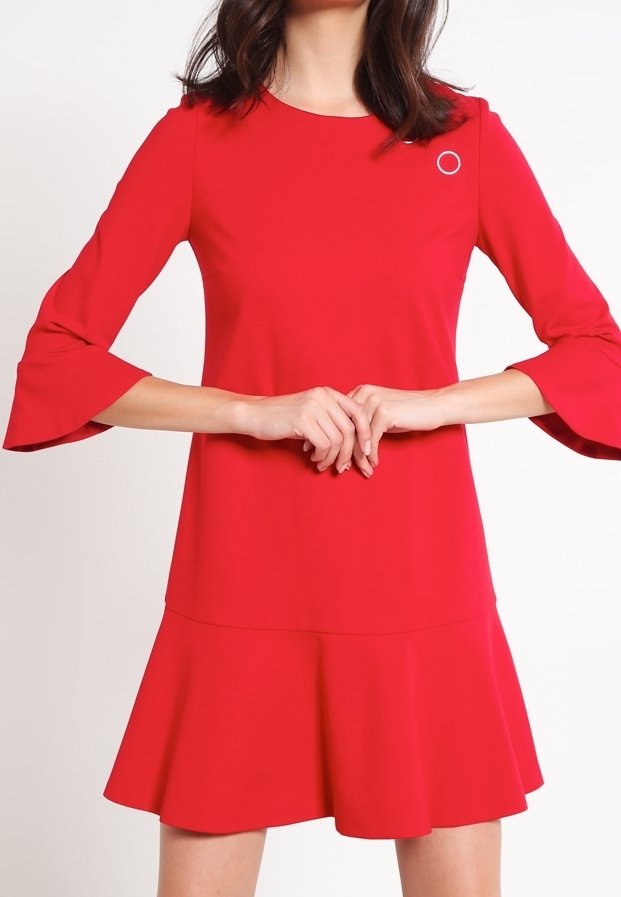 Femme portant une robe rouge vif avec des manches cloche trois-quarts et une jupe évasée, mains jointes devant à la taille.