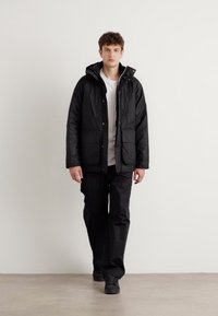 Fekete párnázott dzseki kapucnival, cipzáras záródással és patent gombokkal. Fehér ing fölött viselve, fekete nadrággal és cipővel párosítva.