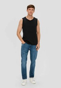 Young man wearing black sleeveless shirt, blue jeans, white sneakers, standing with one hand in pocket against plain background.