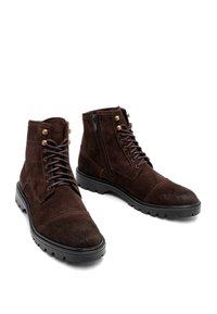 Brązowe zamszowe botki do kostek mają okrągły czubek, przednie sznurowanie i boczny zamek błyskawiczny, złote metalowe akcenty oraz grube czarne gumowe podeszwy.