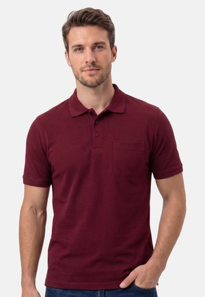 Mann mit kurzen braunen Haaren, der ein bordeauxrotes Poloshirt mit Brusttasche trägt und mit einer Hand in der Tasche vor einem schlichten Hintergrund steht.