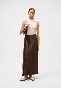 Top sans manches côtelé crème associé à une maxi jupe en satin brun brillant. Le mannequin tient un sac à main métallique et porte des chaussures beiges à clous.