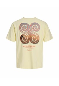T-shirt crème avec un grand motif en spirale orange et gris dans le dos, texte « Sur la Côte d'Azur French Riviera » et une signature sous le motif.