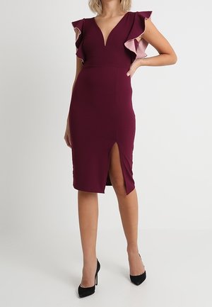 Robe bordeaux M avec un décolleté en V profond, des manches volantées dans une teinte plus claire, une coupe ajustée, et une fente haute sur la cuisse, associée à des escarpins noirs.