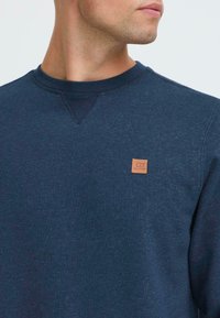 Egy férfi tengerészkék kereknyakú pulóvert visel, amelyen egy kis barna négyzet alakú logó folt látható a bal mellkasán, az oldala felé néz.